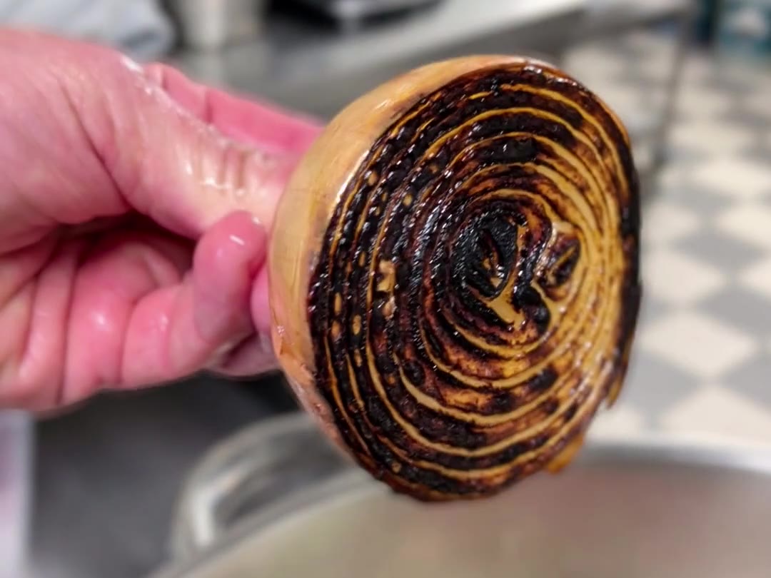 Hand hält eine halbierte Zwiebel mit dunkel angerösteter Schnittfläche über einen Topf mit Gemüsebrühe